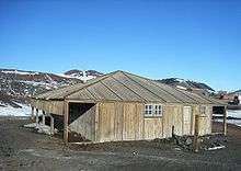  Wooden structure with door and two small windows. To the left is an open lean-to. In the background are partly snow-covered mountains