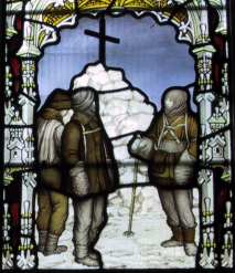 Three figures are depicted in coloured glass, standing by a cairn of snow topped by a large cross. The scene is framed by a decorative arch.