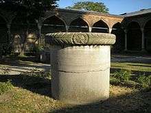 A stout cylindrical column in a courtyard in front of palatial arches of Islamic style