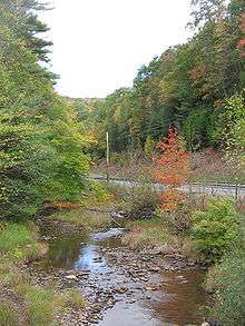 A shallow stream flows over many rocks with a two-lane highway at right. The surrounding woods are starting to show autumn colors, one small tree is bright orange.