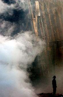 The very small silhouette of a firefighter with smoke in foreground and part of a collapsed building behind him