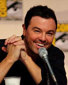 A man with black short hair and a black shirt, with tan skin, laughs into a microphone while leaning forward.