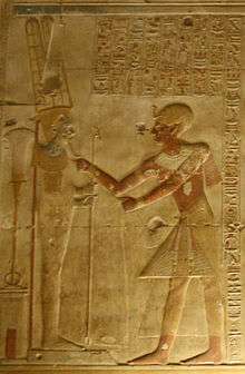 Relief showing an ornately dressed Egyptian man reaching toward a male figure on a pedestal.