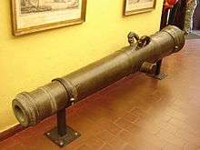 A metal cannon barrel mounted upon two brackets screwed into a brown tiled floor. The cannon barrel is mounted in front of a pale yellow wall supporting two picture frames.