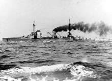 A large gray battlecruiser steams through choppy seas, thick black smoke pours from her rear smoke stack.
