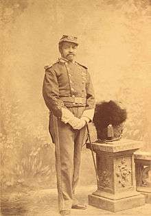 Full-length portrait of a black man with mustache in an ornate military uniform, including white gloves, shoulder boards, cap, and a thin sword tied at his waist. Next to him is a pedestal with a ceremonial military helmet atop.
