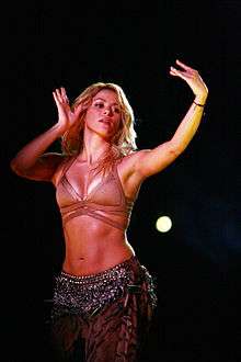 A woman with long blonde hair is belly dancing while wearing a skin-toned halter top which exposes her midriff and a long skirt with glittery fringes.