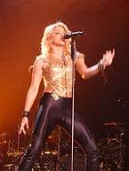 A lady is singing a song dressed in a mesh gold crop top coupled with skin-tight leather pants. She is singing in front of a mic stand.