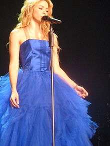 A woman with long blonde hair is singing in front of mic stand while dressed in a feathery blue gown.