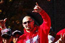 A man in a red hooded sweatshirt and sunglasses raises his hand over his head, showing the "hang loose" sign (making a fist with the thumb and pinky raised).