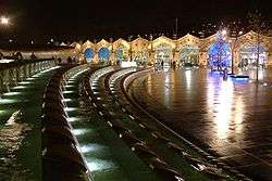 Night view across an open plaza dominated by a long curving water feature that is decoratively lit. At the far side of the plaza there is the arched frontage of a railway station building