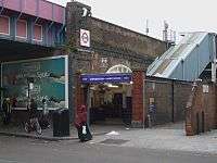 A railway on a brick viaduct crosses a road on a steel bridge, with an entrance below a blue sign reading "SHEPHERD'S BUSH MARKET STATION" in white letters