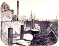 A photograph with two steamships resting in a dewatered drydock with a building housing the engine for operating the lock's gates in the background