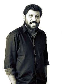 upper-body photo of smiling man with dark hair, moustache and beard wearing dark-colored button down shirt