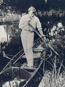 Paget standing in a canoe wearing a deerstalker cap, holding an oar