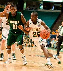 A man in a white jersey with green "SIENA" and "25" on front dribbles a basketball past another man in a forest-green jersey with white "LOYOLA" and "31" on front.