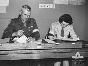 Two soldiers, one male, one female, sit side by side doing paper work at a desk. They are wearing brassards and the sign behind them says: "signal master".