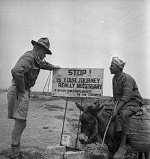A black and white photograph of a man in military uniform and hat inspecting a sign with a man in arab clothing riding a donkey watching on; the sign reads "Stop! Is your journey really necessary? If so give our compliments to the Fuhrer
