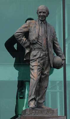 A bronze statue of a bald man wearing a suit. His right hand is on his right hip and he is holding a football to his left hip.