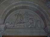Carved stone tympanum showing a bale of cotton and two African slaves overseen by a port official leaning against a desk.