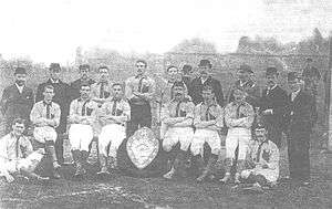 A faded photograph of a posed group of men, outdoors. At the front are six men sitting on benches and two seated on the ground, each dressed in sports clothing, a light-coloured shirt with darker trim, white shorts, dark socks and boots. Standing at the back are eight men, some bearded, wearing hats and coats, and three men in sports clothing. In front of the men is a large trophy in the form of a shield.