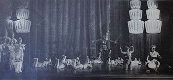 Wide angle shot of the whole stage showing the ballet scene; a threatening character stands at center with a raised sword, while other dancers lie prone on the stage appearing to plead with him; others stand at left looking concerned