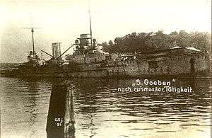 A black and white image of a warship that has become beached. It is listing to starboard. A caption is written in white ink in German over the top of the image.
