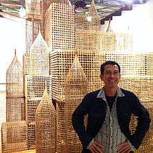 The artist, arms akimbo, in front of a fanciful rattan cityscape.