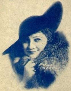 Blue and white picture of a smiling dark-haired woman, facing the camera and looking to the right. She wears a dark brimmed hat and a fur coat. Her right hand is holding the fur coat and there's a ring in her little finger. The text "Sophie Tucker" is written on the picture with small white letters.