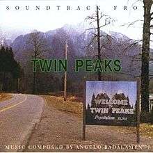 A signpost reading "Welcome to Twin Peaks – Population: 51,201" on a rural roadside, with the main road visible to the left. Several trees (barren and in bloom) and an electricity pole are visible directly behind the signpost; a mountain with snow on its peak is visible in the further background. Brown bold text with a green outline reads "Twin Peaks" in the center; black bold serif text above reads "Soundtrack from"; white bold serif text below reads "Music composed by Angelo Badalamenti".