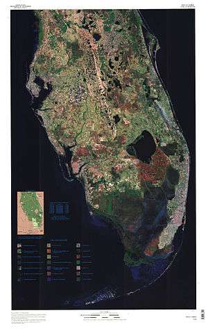 A color satellite image of the lower two thirds of the Florida peninsula: large bodies of water are black, most of the peninsula is spotted green and brown indicating developed and wooded areas; a large chunk of land south of Lake Okeechobee is red, indicating the Everglades Agricultural Area; south of that is a solid swath of dark blue indicating where the Everglades flow in a southwesterly direction into the Gulf of Mexico