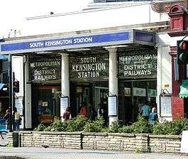 A entrance behind a raised bed of green plant and under a blue sign reading "SOUTH KENSINGTON STATION" leads into an arcade of shops. Above the entrance there is a glass panel with white lettering reading "METROPOLITAN AND DISTRICT RAILWAYS" and "SOUTH KENSINGTON STATION".