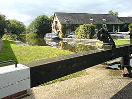 A canal lock in the sunshine.  Behind the lock is a Victorian brick building, formerly a pumping station, now a private residence.