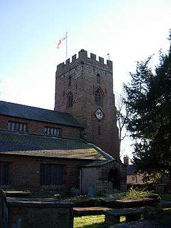 The tower and part of the body of a church seen from the northeast. On the tower is a flag and on its north face is a clock; tombs are in the foreground