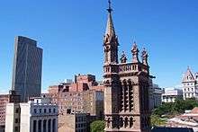 A closeup view of the upper section of the church's tower, with a cityscape behind it. A tall modernistic building is at left; a tower with a red peaked roof is at right