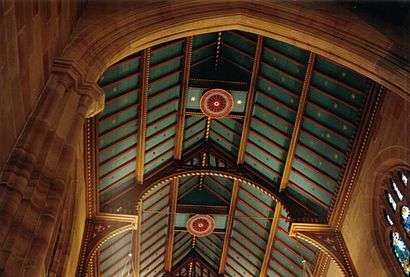 View looking up into the open gabled roof of St Andrews. The beams, rafters and air vents are decorated with bright red rebates and gold leaf details. The ceiling panelling is blue-green dotted with gold stars.