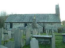 A very simple, small church seen from the north with a small bellcote on its right. There are many gravestones and tombs in the foreground