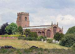 A church seen from the southeast at a distance; the crenellated tower is to the left and the body of the church has Perpendicular style windows. In front of the church is a field with bushes to the left and right