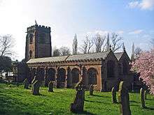 A substantial red sandstone church seen from the southeast. The tower and body of the church are crenellated.  In the churchyard are gravestones and at the right extremity is a tree in flower