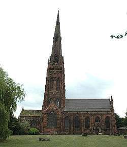 A sandstone church with a very tall spire and a protruding transept seen from the south. In front of the church is a grassed area containing a bench and to the left is a tree