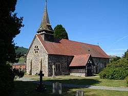 A church built in irregular stones with a red tiled roof seen from the southwest. On the west front is a round window and above this is a small spire.  Protruding from the south wall is a red-tiled, timber-framed porch