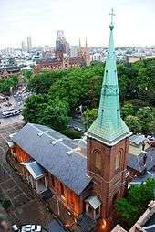 The church is seen from above, showing the spire and slate roof. The nearby park is full of dark leafy trees. In the distance is a large sandstone cathedral.