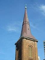 A detail of the tower and spire showing that it is octagonal and shaped rather like a witch's hat. In this view the copper has been renewed and appears brown and lustrous. On top are a cross and ball with green patina. The tower is brick, has flat corner pilasters and large louvred windows.