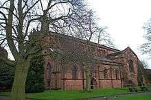 A red sandstone church seen from the northeast, with a clerestory and a gabled transept. To the left and in front of the church are leafless trees