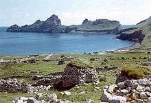 A rocky bay with stone ruins in the foreground. The ocean enters the picture from the left and across the bay several rocky crags are visible sticking out the sea.