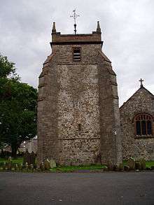 A broad windowless tower with angle buttresses; to the right is the west wall of the nave; on the left is a tree