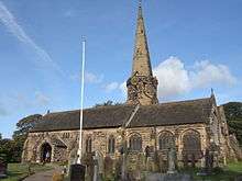 A stone church with a central round tower with clock faces surmounted by an octagonal spire.