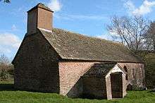 A small, simple, stone church with a stone slate roof, a bellcote with a pyramidal roof, a porch and small windows, seen from the southwest