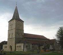Three-quarter view of a flint church with a tall, three-stage tower to the left, topped with a dark grey spire.  Low projections with paired arched windows flank the tower on both sides.  A porch juts forward from the main body of the church, next to three low lancet windows.  The roofs are of red tiles.  Irregularly spaced gravestones are in front of the church.