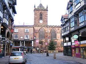 In the centre is a church tower with a small pyramidal spire; to the right is the body of the church and to the left a building containing a shop and part of Chester Rows. In front of the church is a street on each side of which are black-and-white buildings containing shops and Rows, with the back of a grey-coloured car in the left foreground